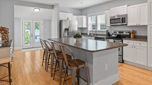 Une cuisine moderne avec des meubles blancs, un grand îlot central doté de tabourets, des appareils en acier inoxydable et une vue sur l’extérieur.