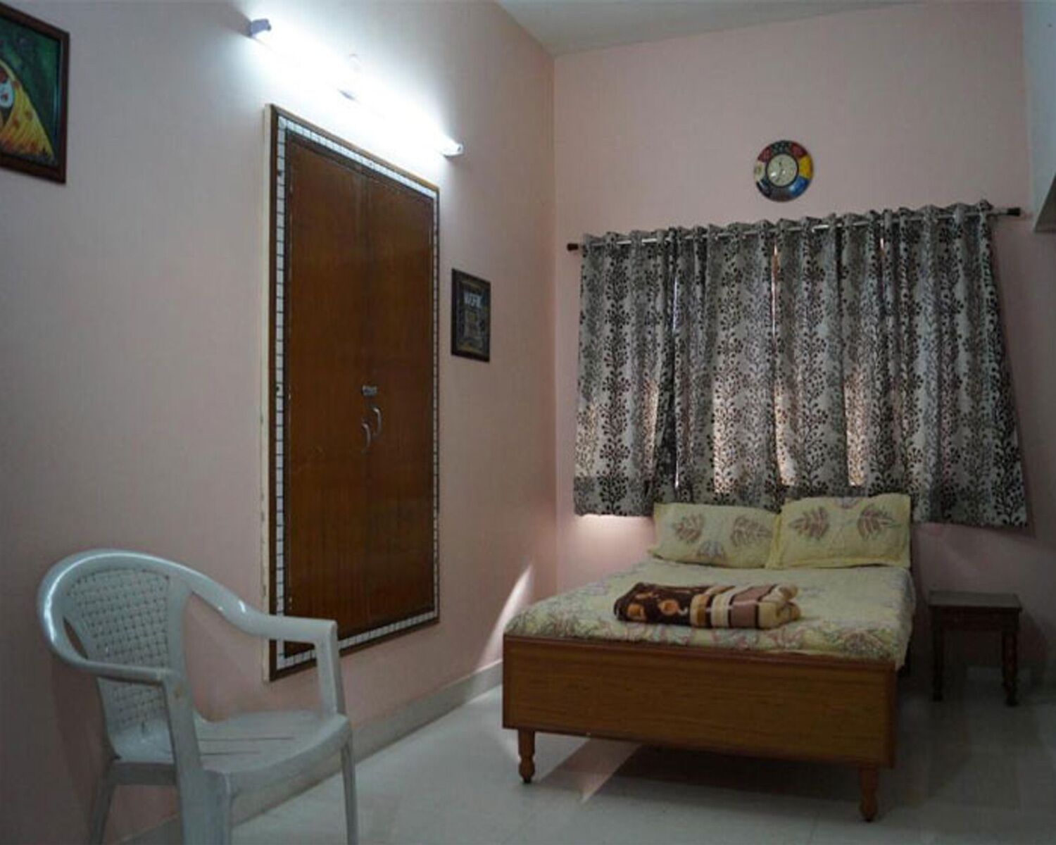 Un dormitorio con una cama de madera, una silla de plástico blanco, un armario de madera, cortinas estampadas y un reloj de pared.