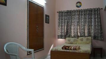 Un dormitorio con una cama de madera, una silla de plástico blanco, un armario de madera, cortinas estampadas y un reloj de pared.