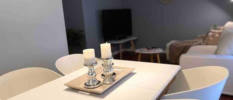 A modern living room with a dining table set for two, featuring white chairs and a tray with candles.