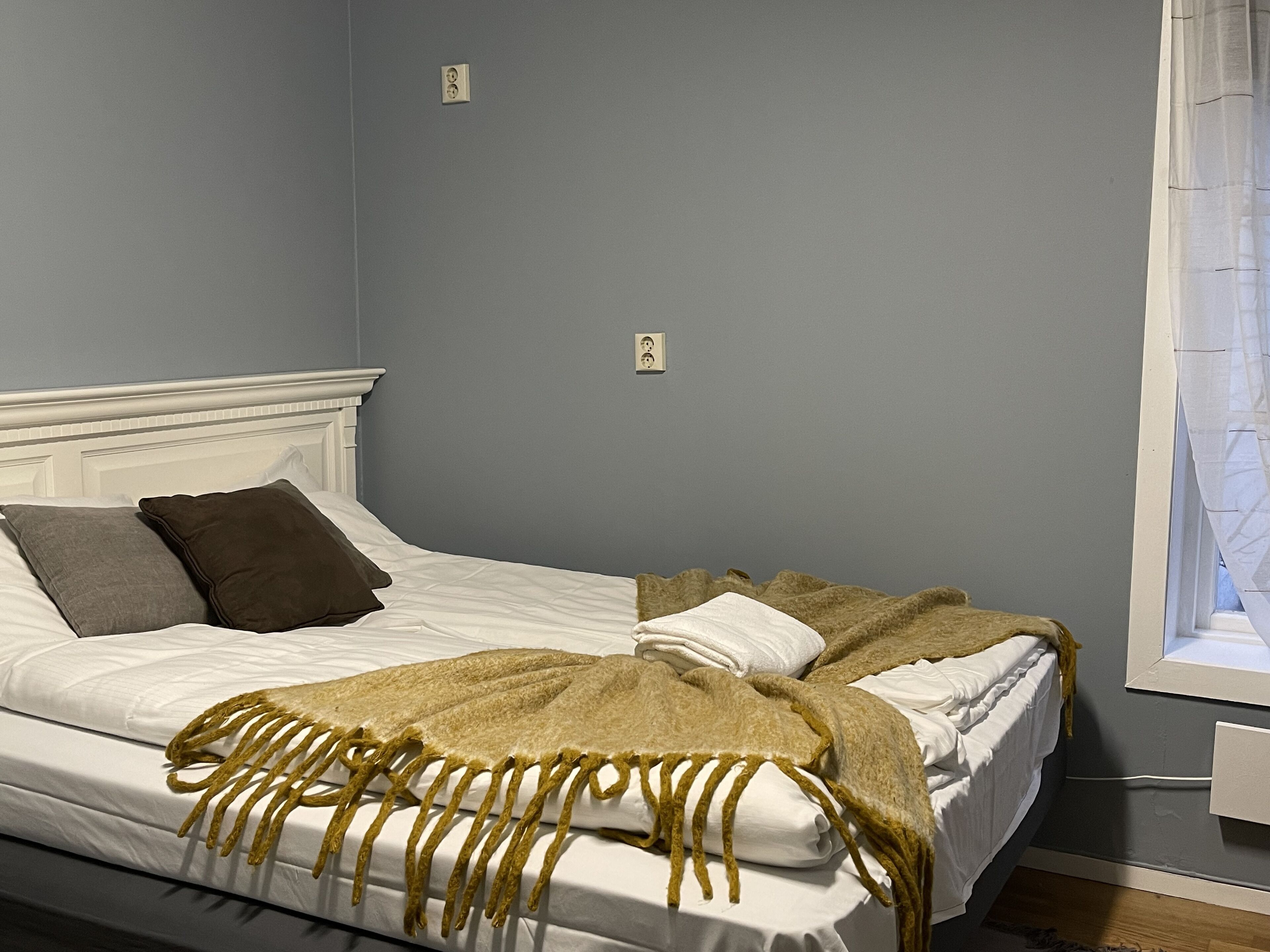 A bed with white bedding and a brown throw blanket, a white headboard, and a window with a curtain.