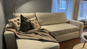 A beige sofa with a throw blanket and pillows, a floor lamp, and a view of a window with blinds. - Charming 2-Bed Apartment in Stavanger (Stavanger)