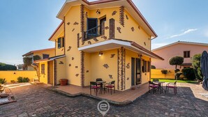 A two-story house with a yellow facade, a balcony with black railings, and a paved patio area with outdoor seating. - Villa Monica - Iglesias (Iglesias)