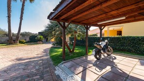 A covered outdoor area with a paved walkway, palm trees, and a parked motorcycle. - Villa Monica - Iglesias (Iglesias)