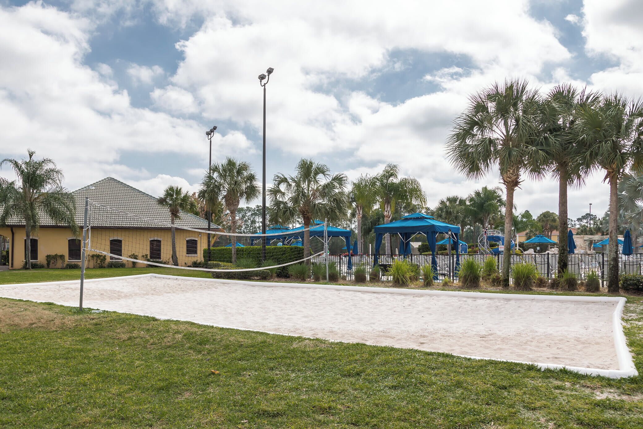 A beach volleyball court with a net, surrounded by palm trees and a building with a tiled roof.