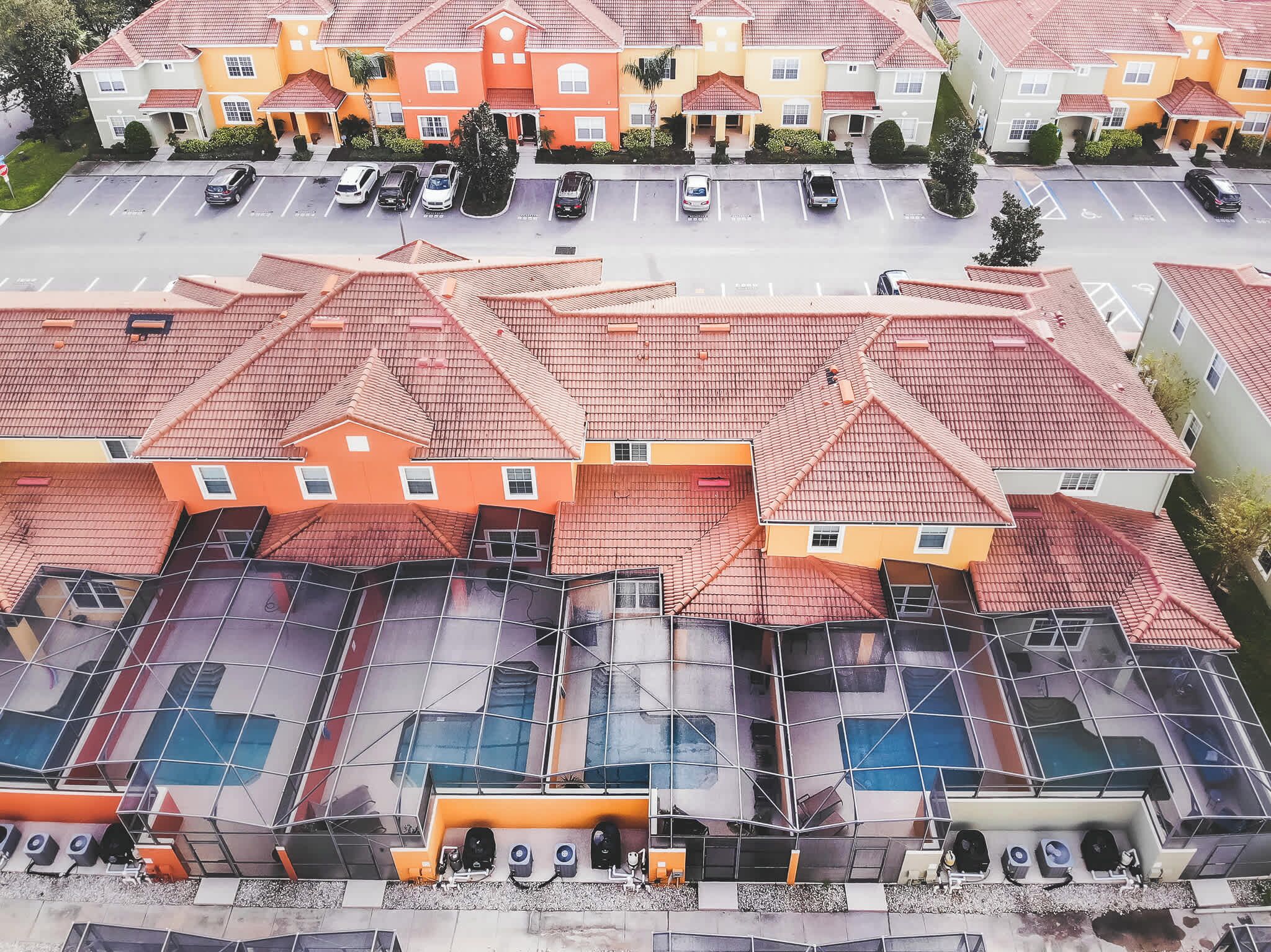 A residential area with multiple houses, each with a red-tiled roof and orange walls. There are several cars parked in the driveway, and a swimming pool is visible on the ground floor of one of the houses.