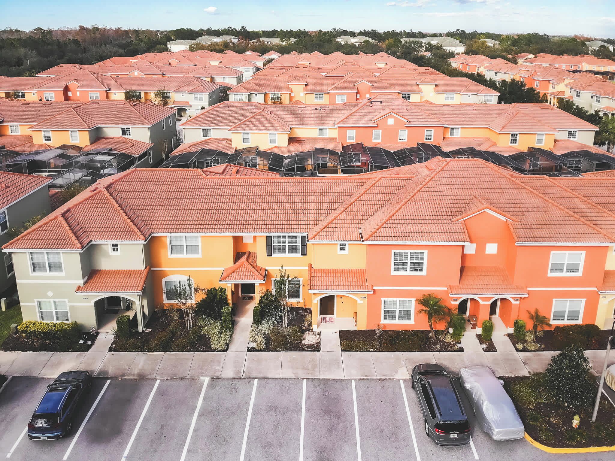 A residential area with multiple two-story houses, each with a red-tiled roof and a mix of beige and yellow exteriors. There are parking spaces in front of the houses.