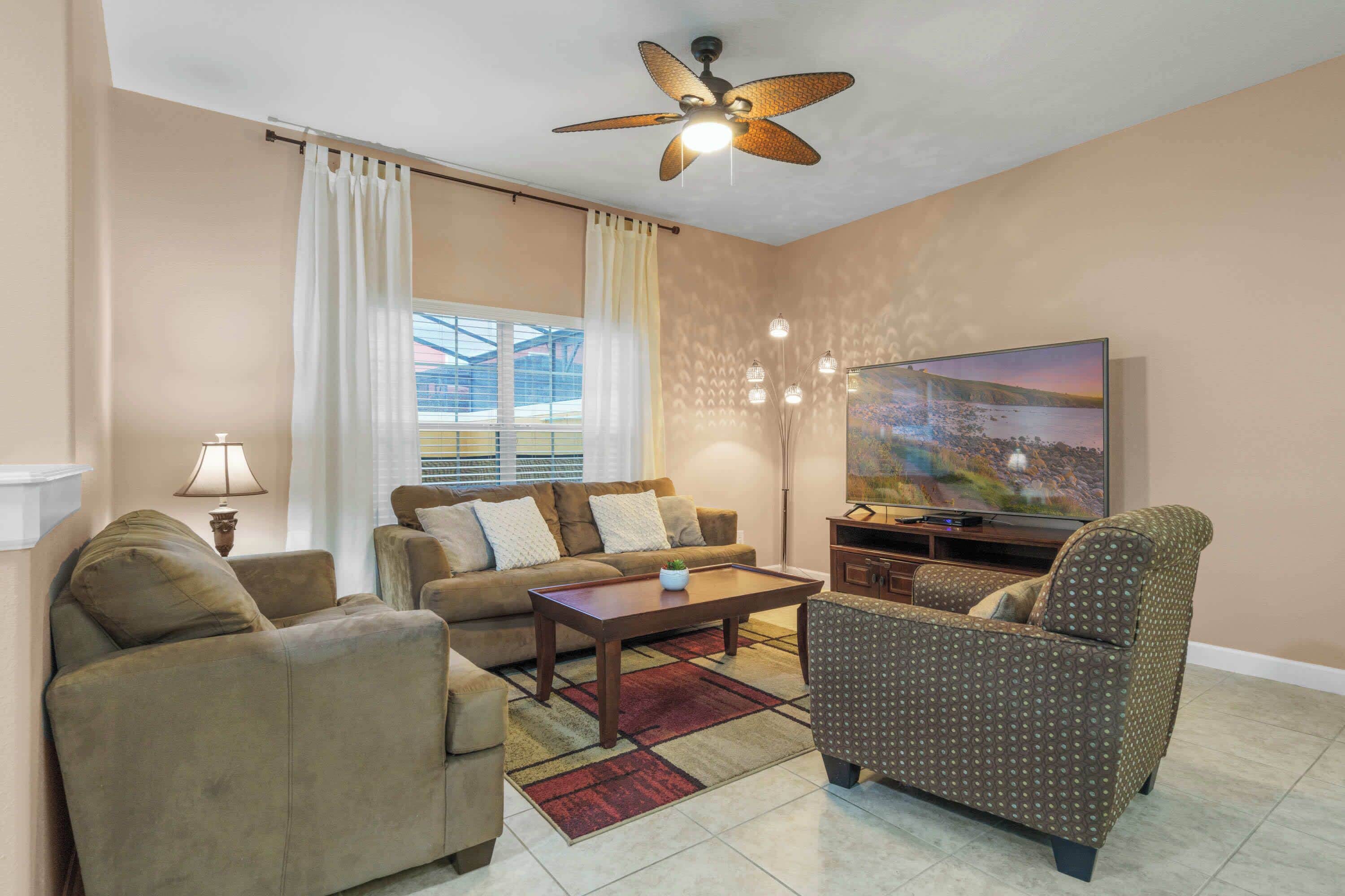 Uma sala de estar com sofá, poltrona, mesa de centro, TV e ventilador de teto.