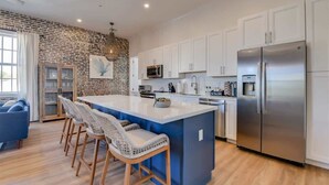 Una cocina moderna con una isla grande, armarios blancos, un refrigerador de acero inoxidable y una pared decorativa de ladrillo.