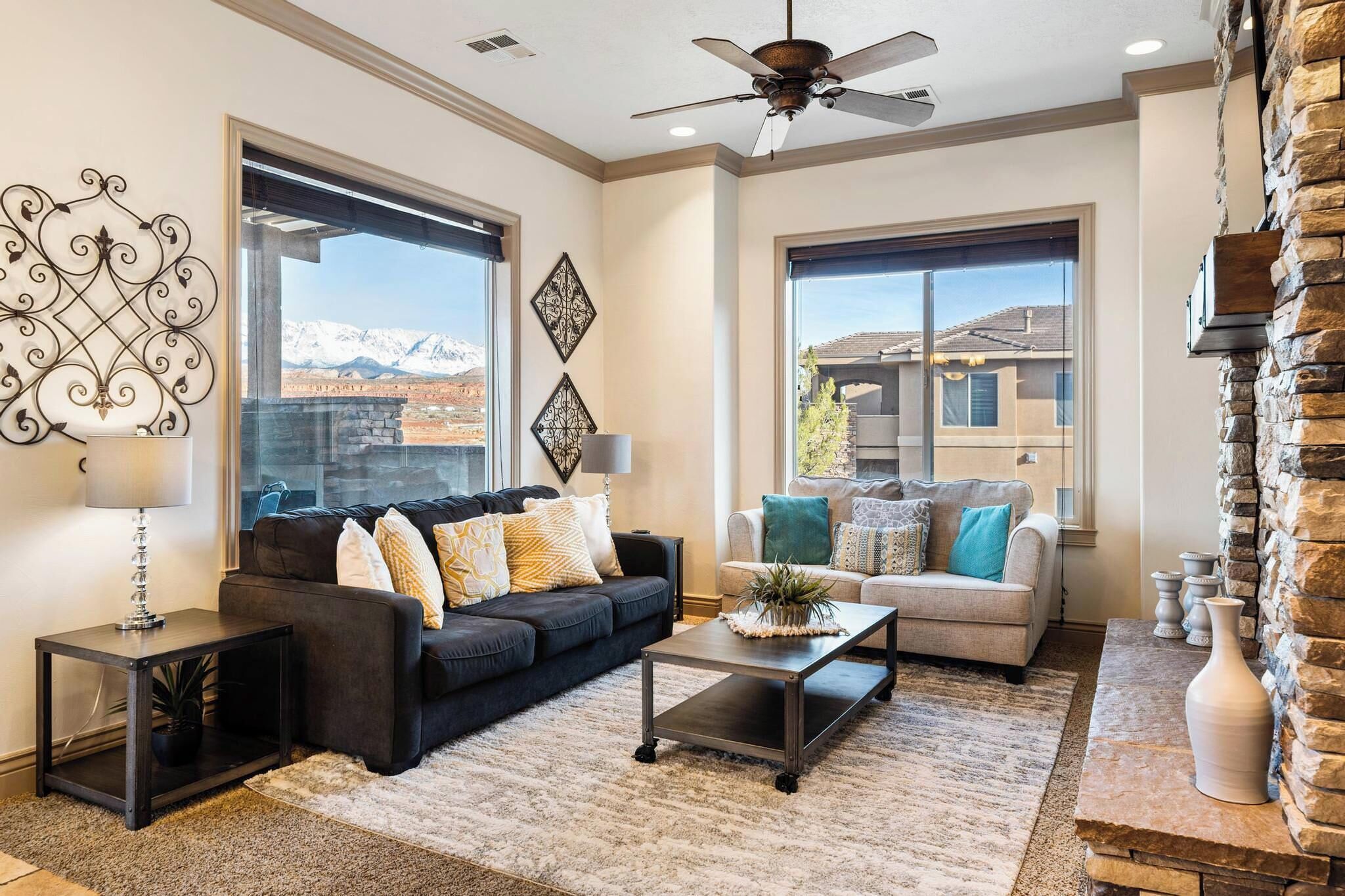 A spacious living room with a dark sofa, a light-colored sofa, and a coffee table. There is a large window with a mountain view and a ceiling fan.