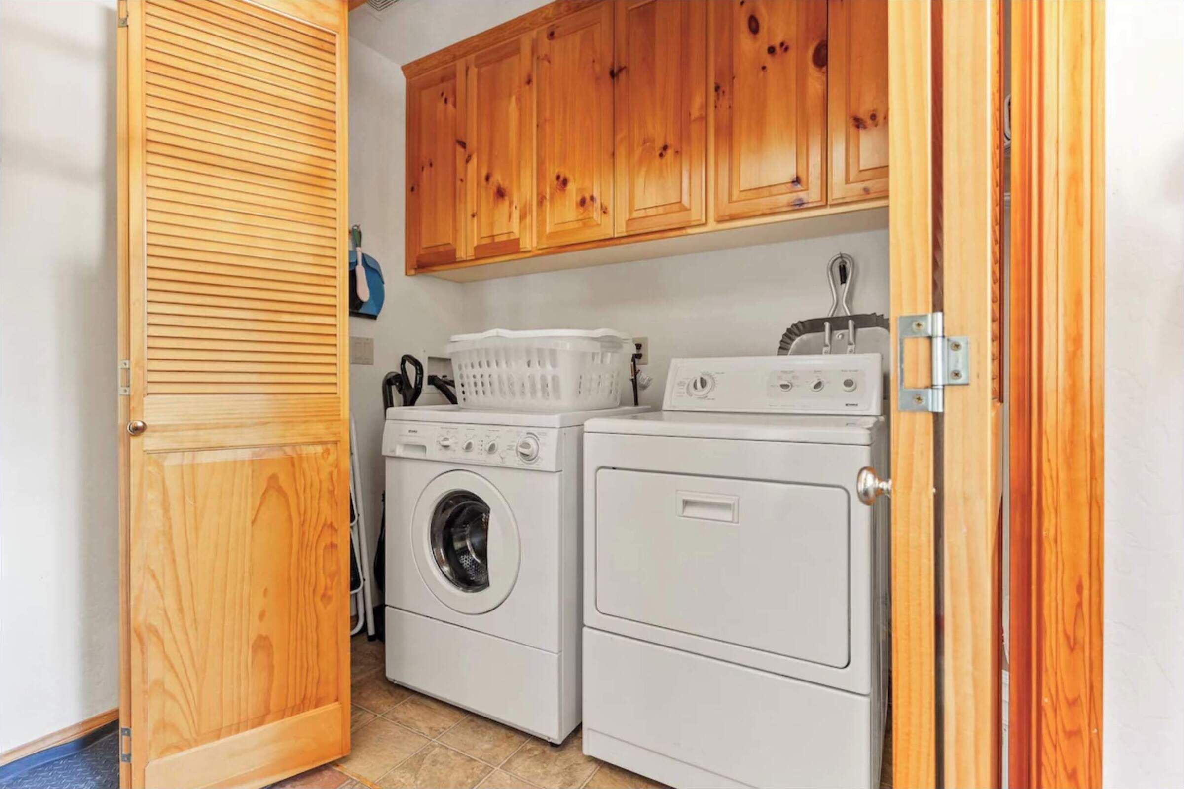 A laundry room with two washing machines and a dryer, wooden cabinets above, and a door with a louvered design.