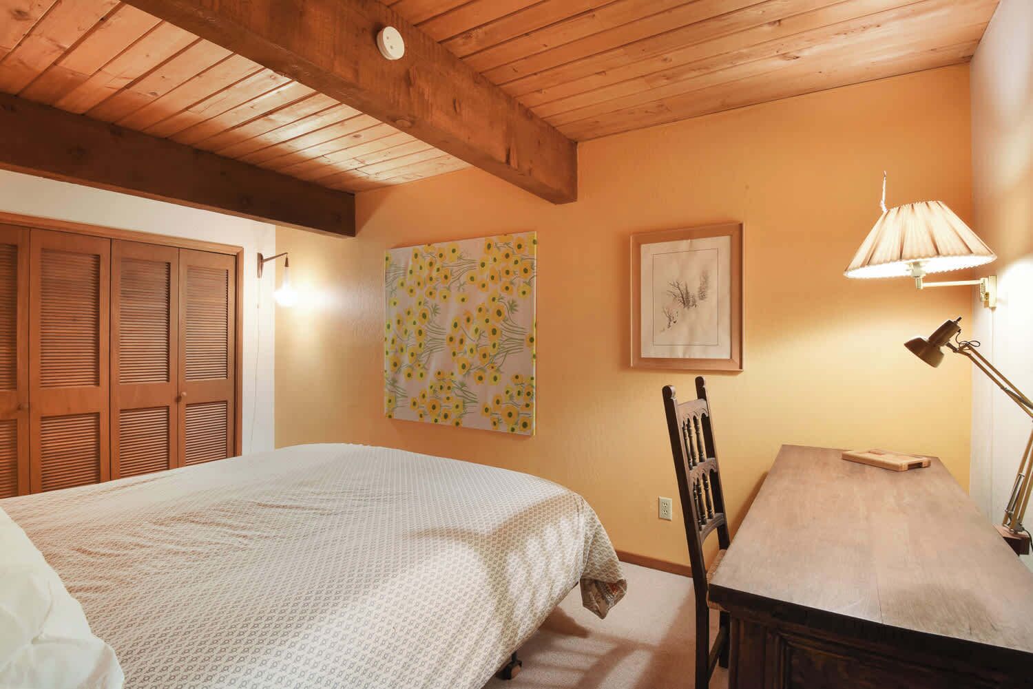 A bedroom with a bed, a wooden ceiling, a desk with a lamp, and a chair.