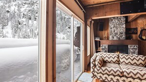 Indretning i en hyggelig hytte med en stenvægspejs, en sofa med mønstrede puder og udsigt til en sneklædt skov gennem store skydedøre.