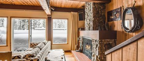 A cozy cabin interior with a stone fireplace, wooden walls, a large window with a snowy view, and a chevron-patterned sofa.