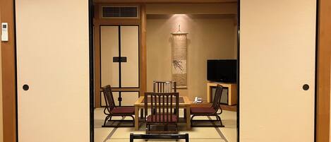 A traditional Japanese room with tatami flooring, a low table with chairs, a television, and sliding doors.