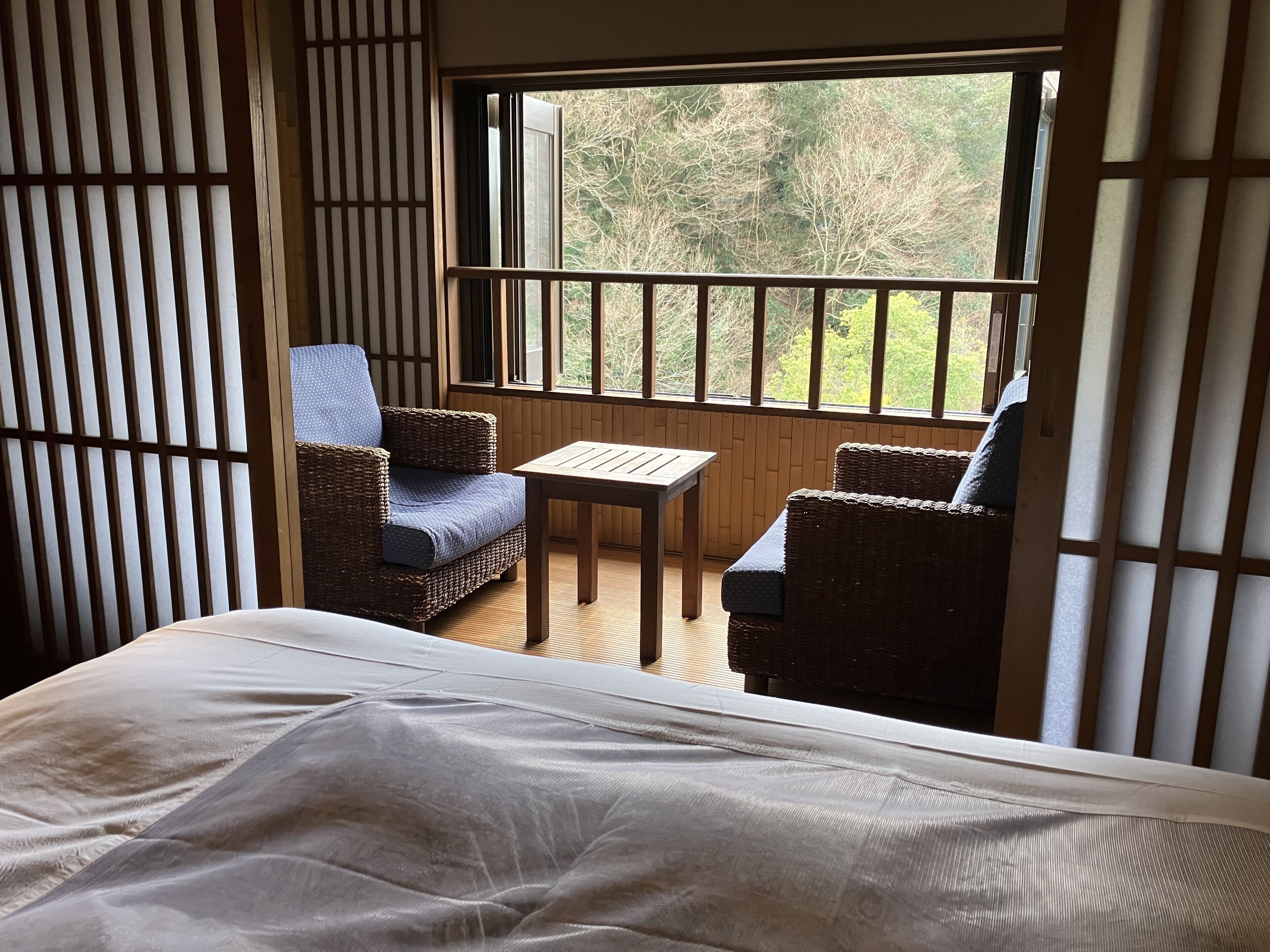 A room with a bed, two wicker chairs, a small table, and a window with a view of trees.
