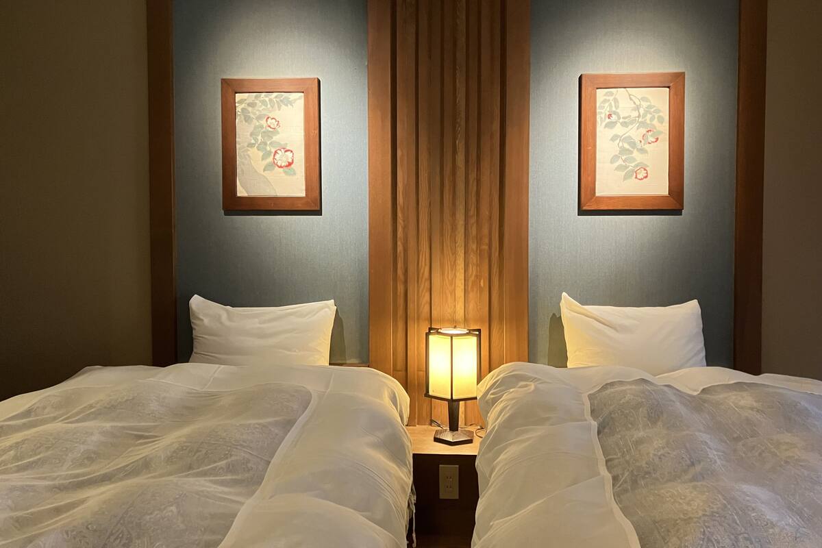 Een hotelkamer met twee bedden, een nachtkastje met een lamp, en twee florale kunstwerken in lijsten aan de muur.