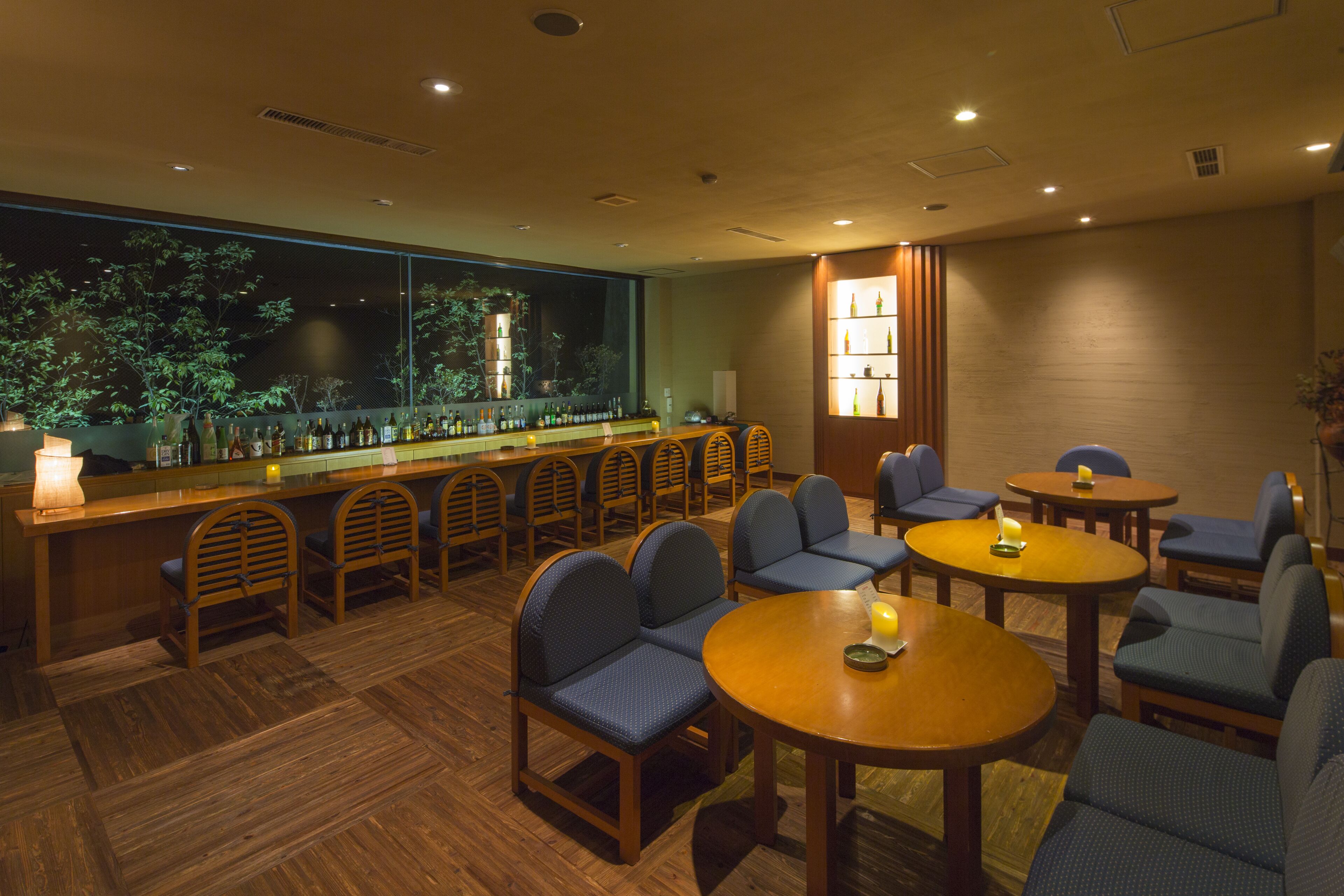 Un área de bar con sillas y mesas de madera, un ventanal con vistas a árboles, y una estantería con botellas.