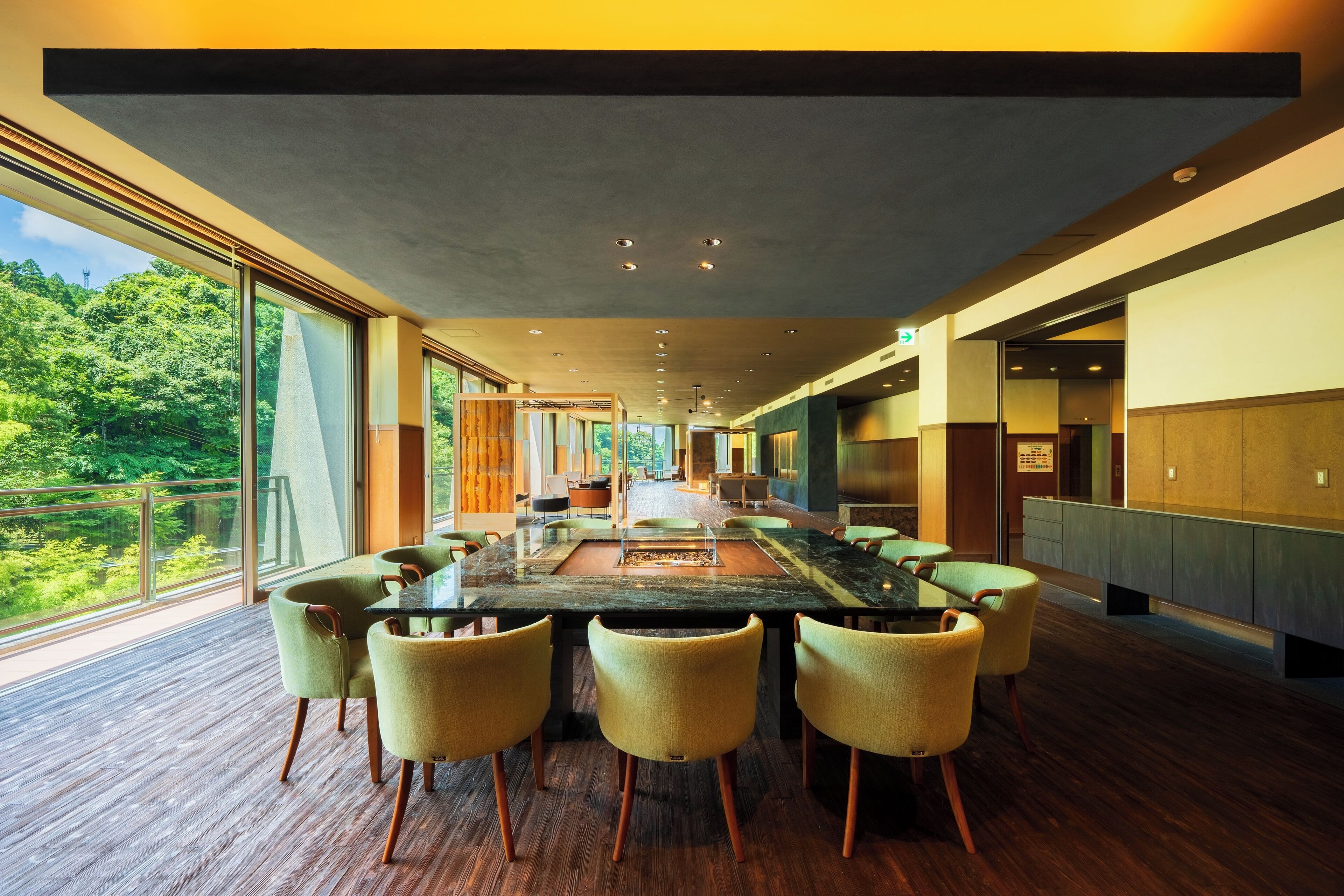 A modern dining area with a long table and chairs, large windows offering a view of the outdoors, and a wooden floor.
