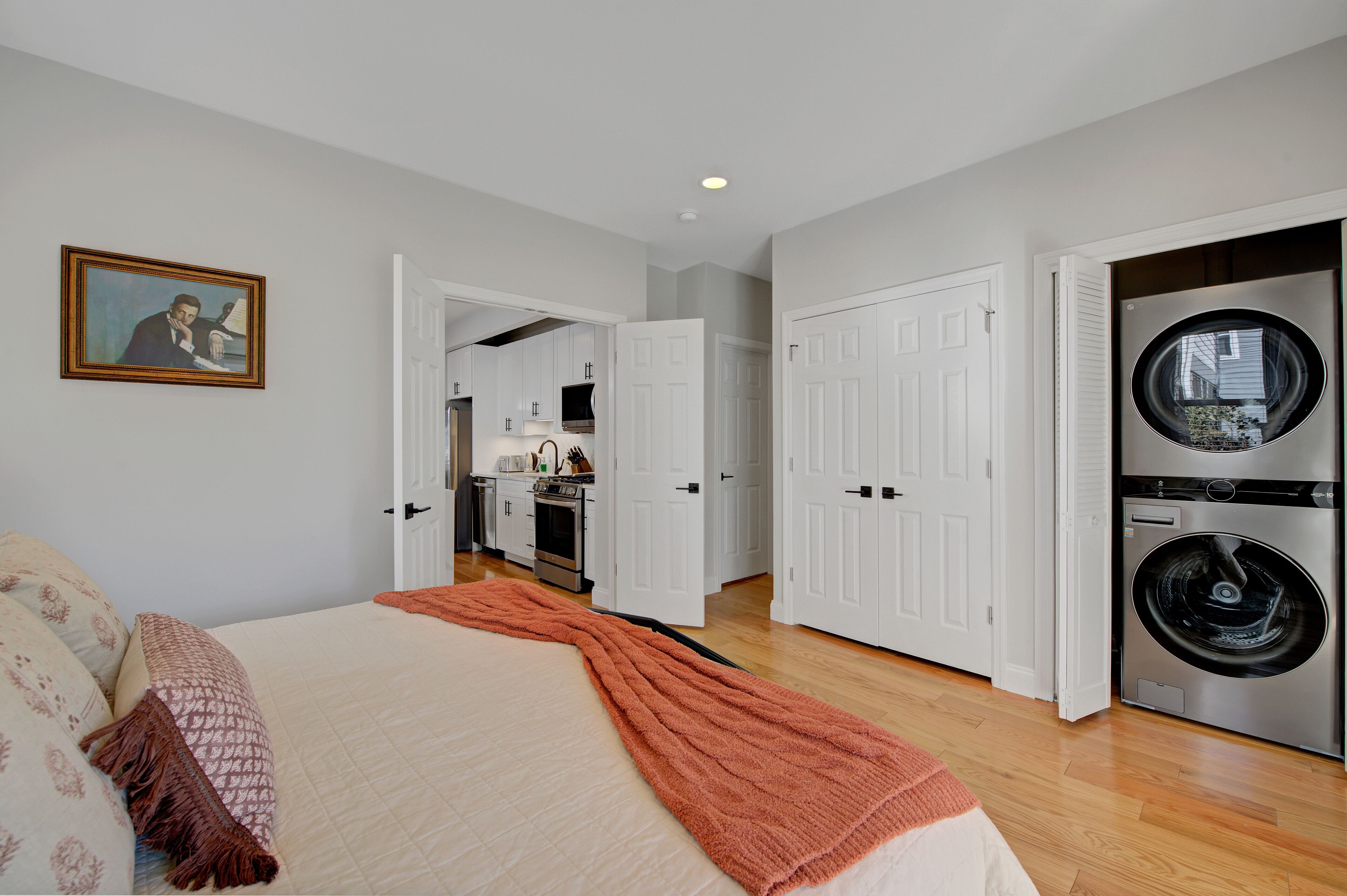 A bedroom with a bed, a painting, and a hallway leading to a kitchen and laundry area.