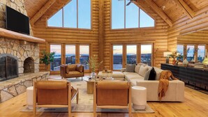 A spacious log cabin interior with a stone fireplace, leather furniture, and large windows offering scenic views.