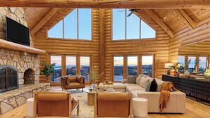 A spacious log cabin interior with a stone fireplace, large windows, and a cozy seating area.