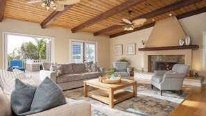 A spacious living room with a high ceiling, wooden beams, a fireplace, and a large window overlooking a balcony.