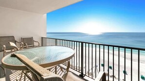 Ein Balkon mit Glastisch und Stühlen, der Blick auf einen Strand mit blauem Himmel und ruhigem Wasser gerichtet ist.