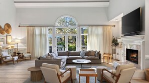 A spacious living room with a large sectional sofa, a fireplace, a television, and large windows with curtains.