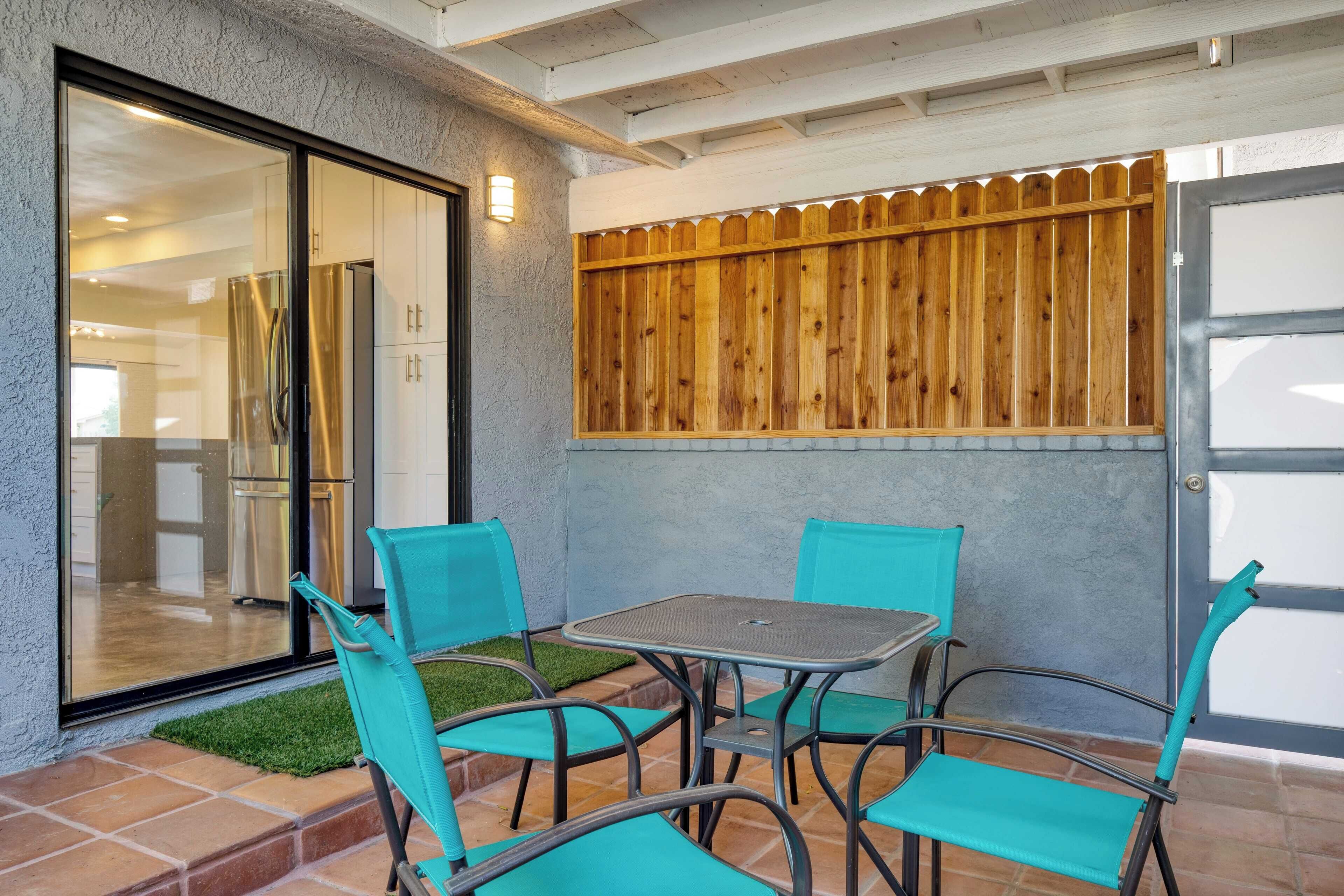 A patio with turquoise chairs and a table, a wooden fence, and a sliding glass door leading to a kitchen.