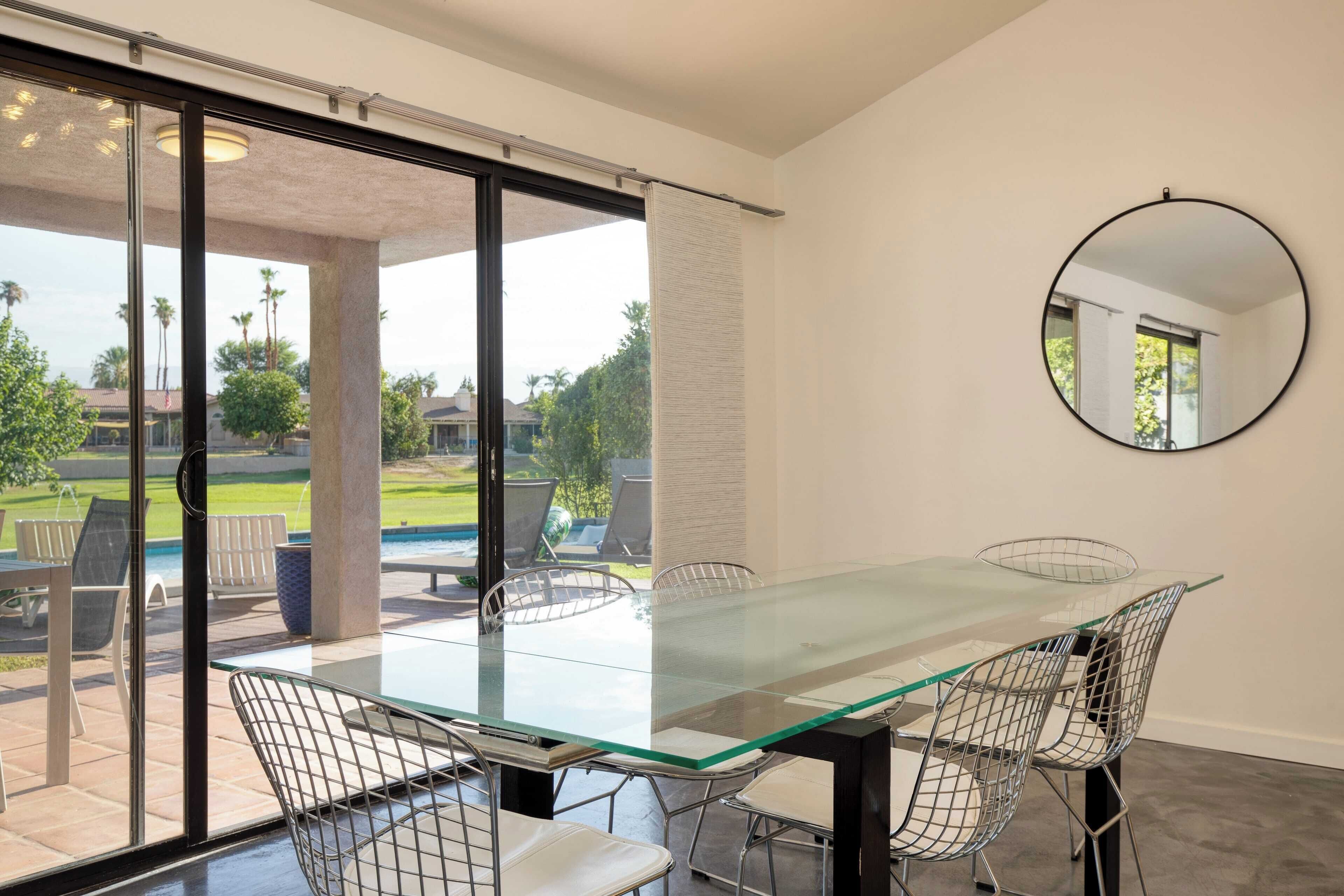 A modern dining area with a glass table and metal chairs, large sliding glass doors leading to an outdoor patio with chairs and a pool.