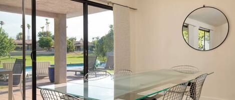 A modern dining area with a glass table and metal chairs, large sliding glass doors leading to an outdoor patio with chairs and a pool.