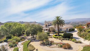 A large, single-story house with a curved driveway, surrounded by lush landscaping and a palm tree. - Modern Temecula Mansion w Pool, Hot Tub, Guest House & Valley Views Camila by Avantstay (Temecula)