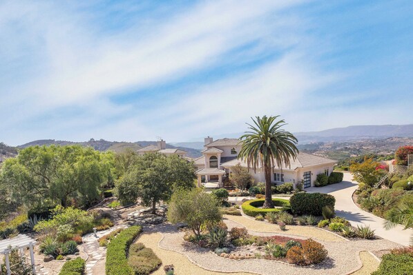 A large, single-story house with a curved driveway, surrounded by lush landscaping and a palm tree. - Modern Temecula Mansion w Pool, Hot Tub, Guest House & Valley Views Camila by Avantstay (Temecula)