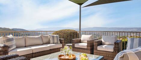A terrace with wicker furniture, a glass table, and a view of distant hills.