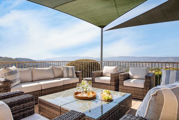 Eine Terrasse mit Korbmöbeln, einem Glastisch und Blick auf ferne Hügel.