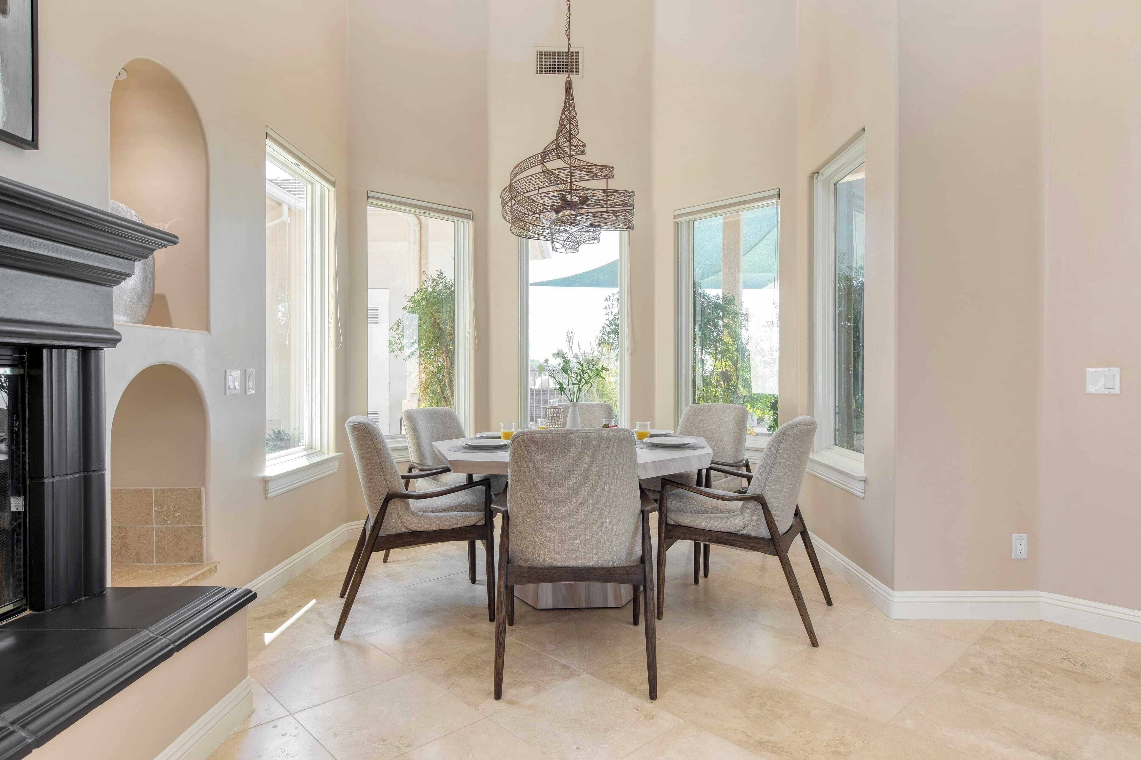 Un comedor con una mesa redonda, seis sillas, una araña y ventanas amplias.
