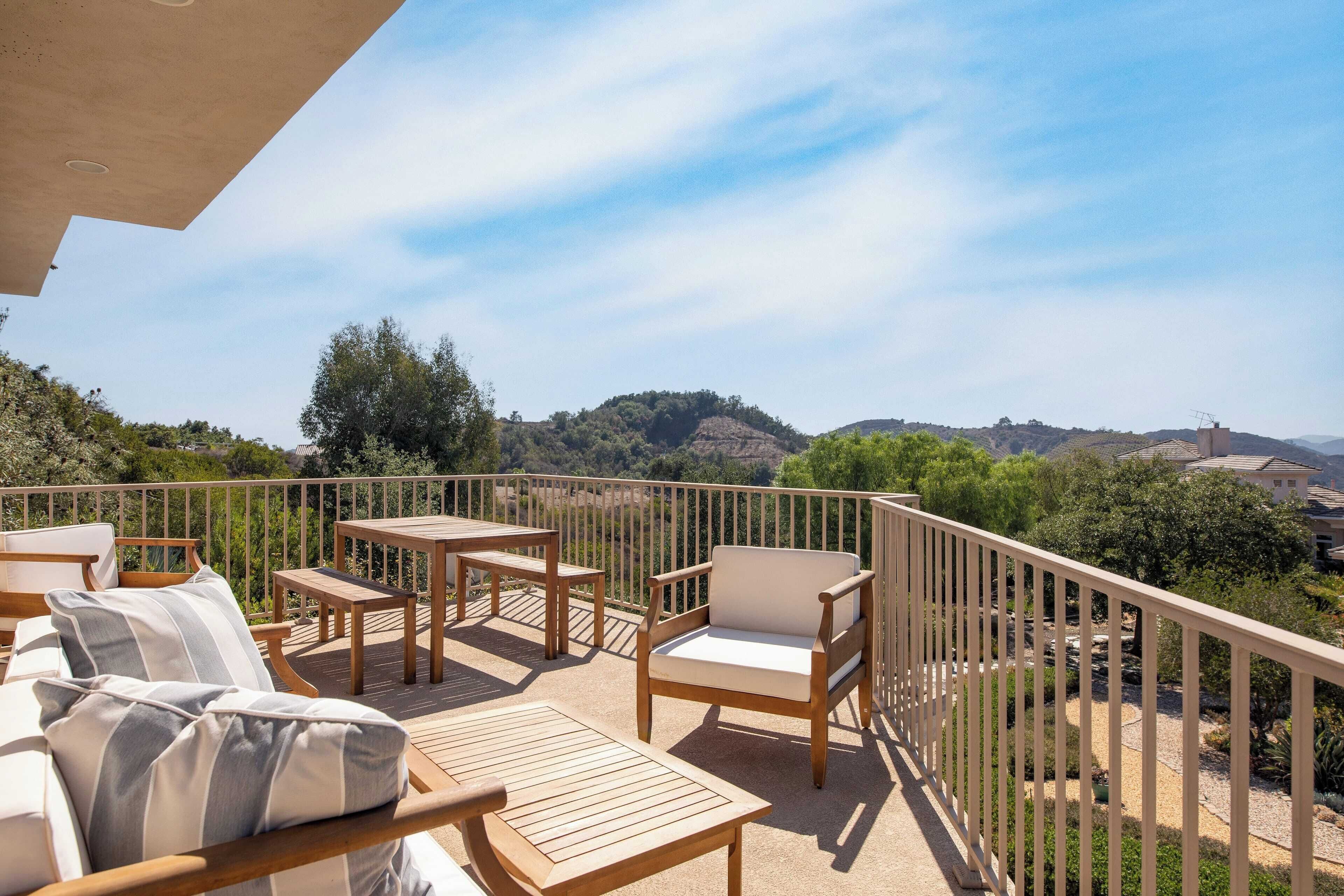 A balcony with wooden furniture, including a sofa, chairs, and tables, overlooking a scenic landscape with hills and trees.