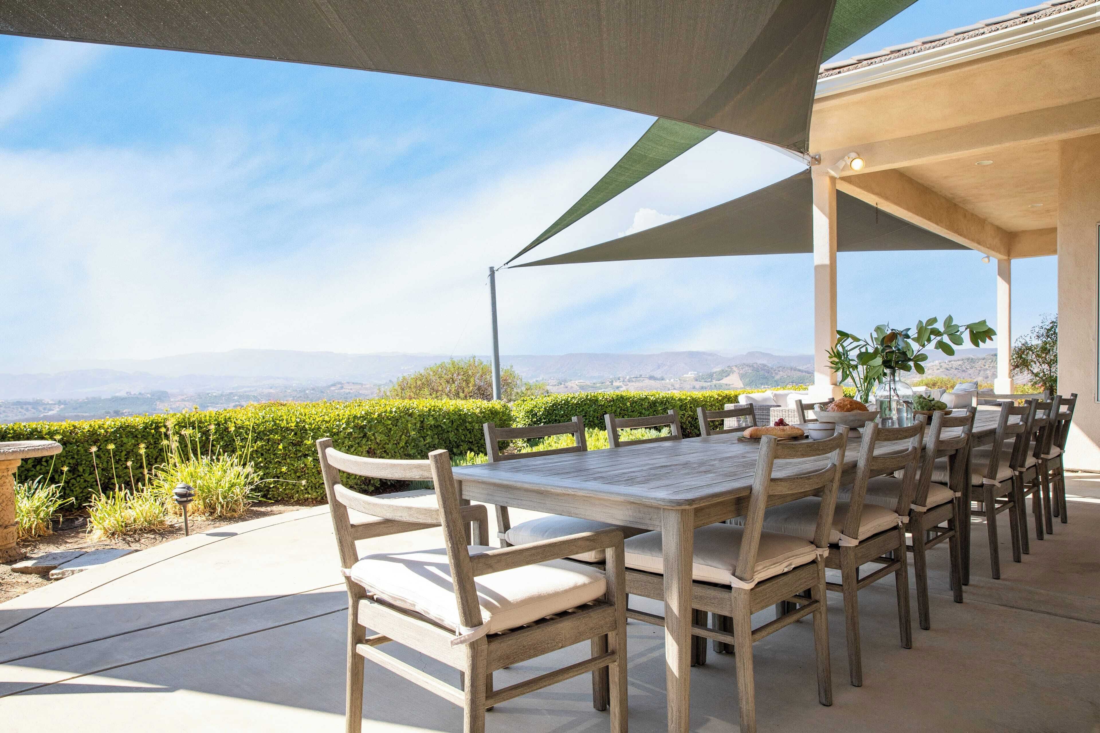 An outdoor patio with a long wooden table and chairs, surrounded by a large canopy, overlooking a scenic landscape with mountains and a clear sky.