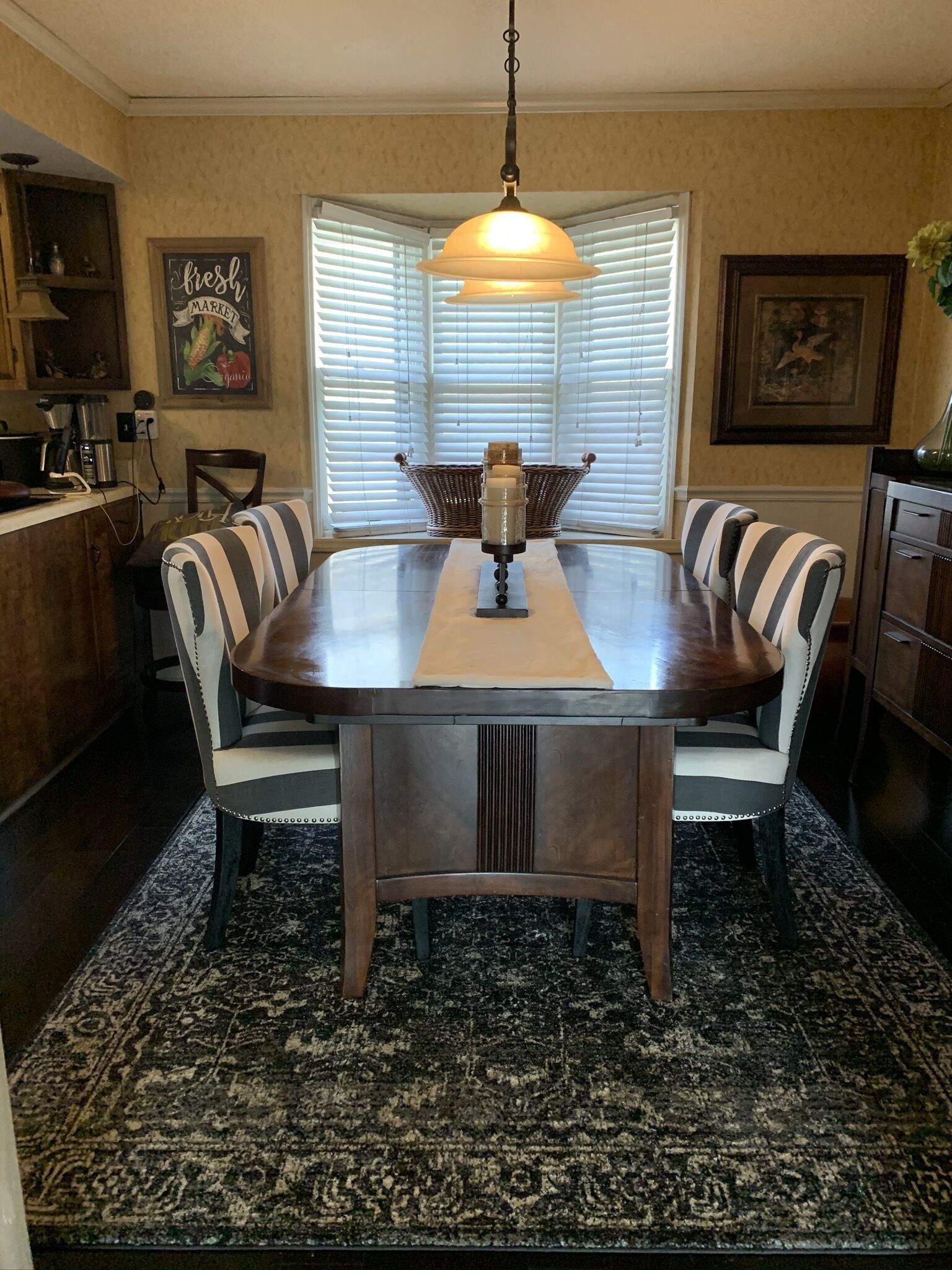 Un coin repas avec une table ronde, des chaises, un luminaire suspendu, une fenêtre avec des stores et, au fond, un espace cuisine.