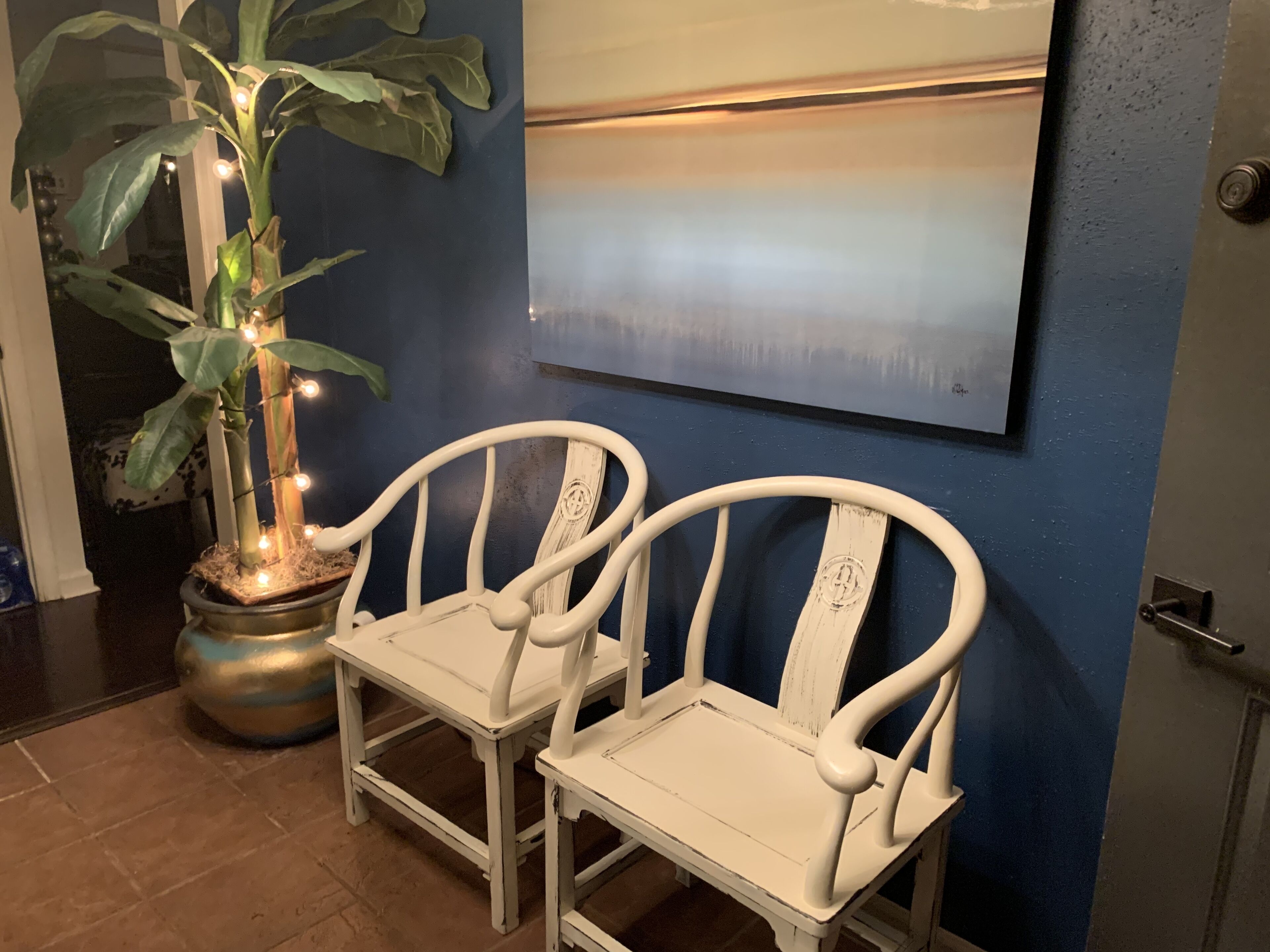 Deux chaises blanches, dotées d’un dossier et d’accoudoirs incurvés, sont placées sur un sol carrelé, à côté d’une plante en pot décorée de guirlandes lumineuses.