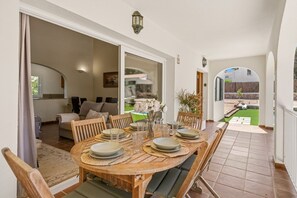 A dining area with a wooden table set for four, a sofa, and a view of a garden through an open door.