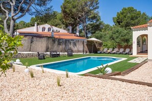 A swimming pool with lounge chairs, a stone wall, and a building with a tiled roof.