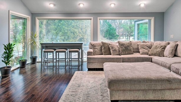 A modern living room with a sectional sofa, a dining area with a small table and chairs, and large windows offering a view of trees. - Spacious Rochester Home w/ Heated Pool & Hot Tub! (Rochester)
