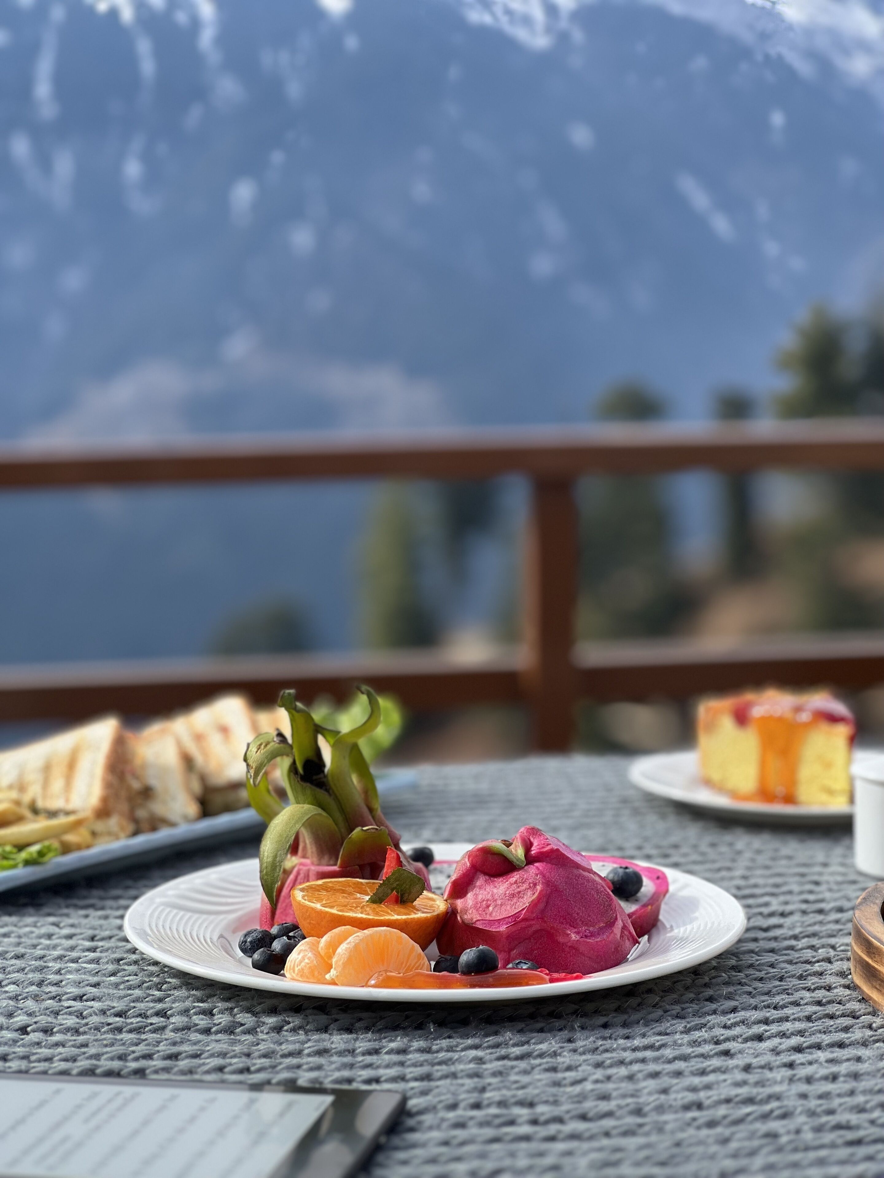 A table with a plate of fruit and a slice of cake, set against a mountain backdrop.