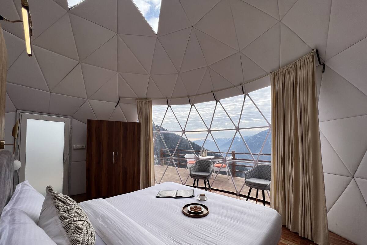 A bedroom with a bed, a wooden wardrobe, a window with curtains, and a view of mountains.
