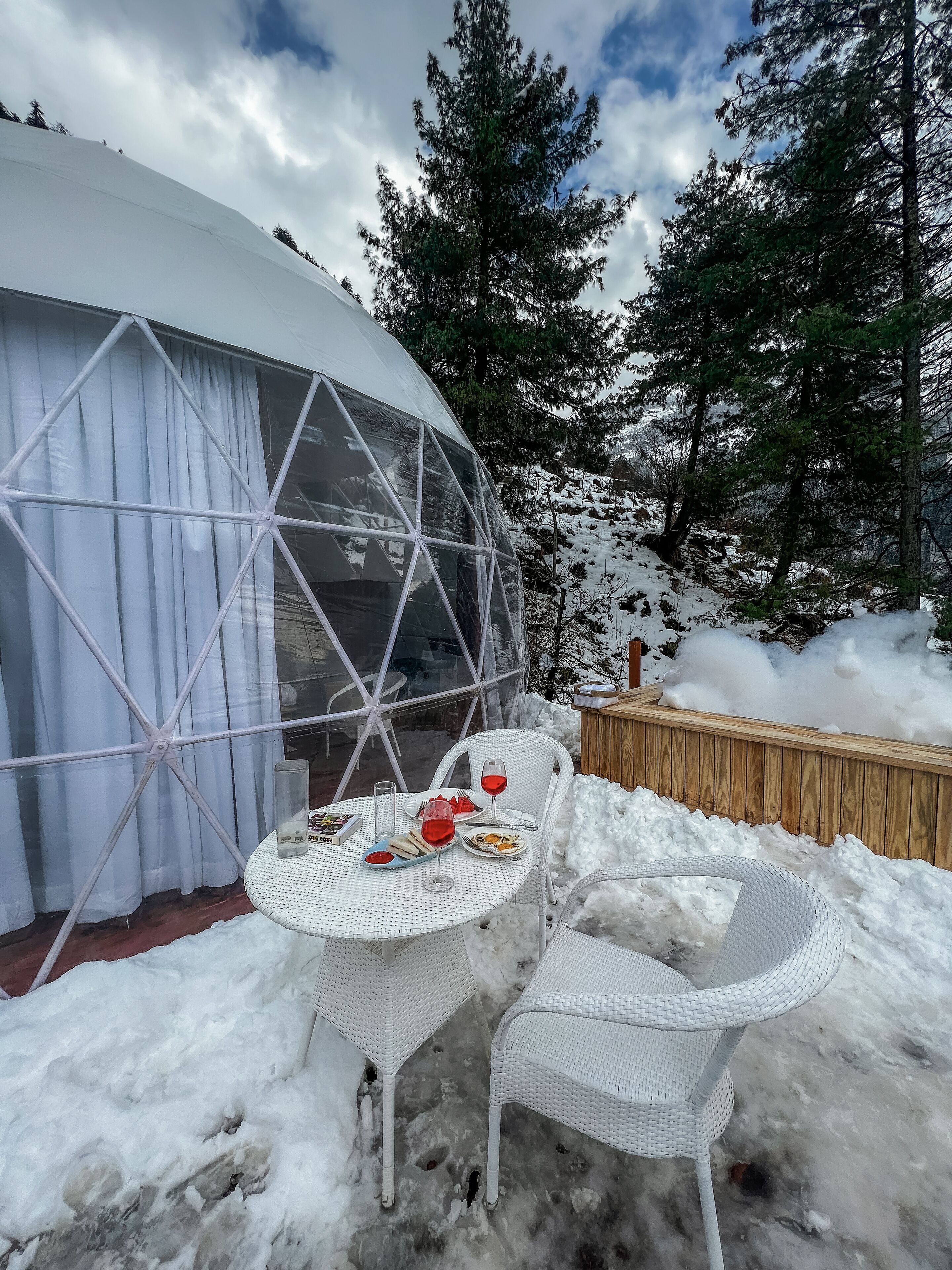 A snow-covered outdoor dining area with a round table and chairs, a glass dome structure, and a wooden deck.