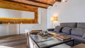 Ein modernes Wohnzimmer mit einem Glastisch, einem grauen Sofa und einer Küchenzeile mit weißen Schränken und einer Ziegelwand.