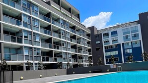 A modern hotel with a pool, featuring multiple balconies and a clear blue sky.