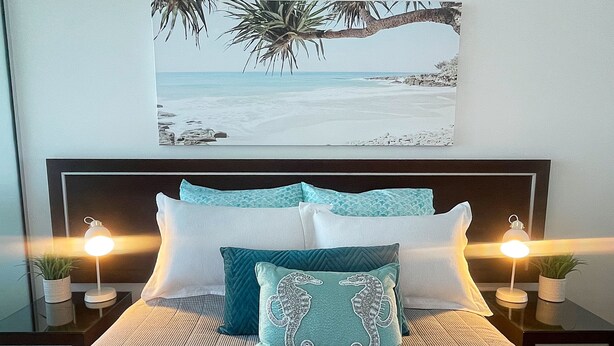 A neatly made bed with a patterned bedspread, two matching pillows, and a framed picture of a beach scene above it.